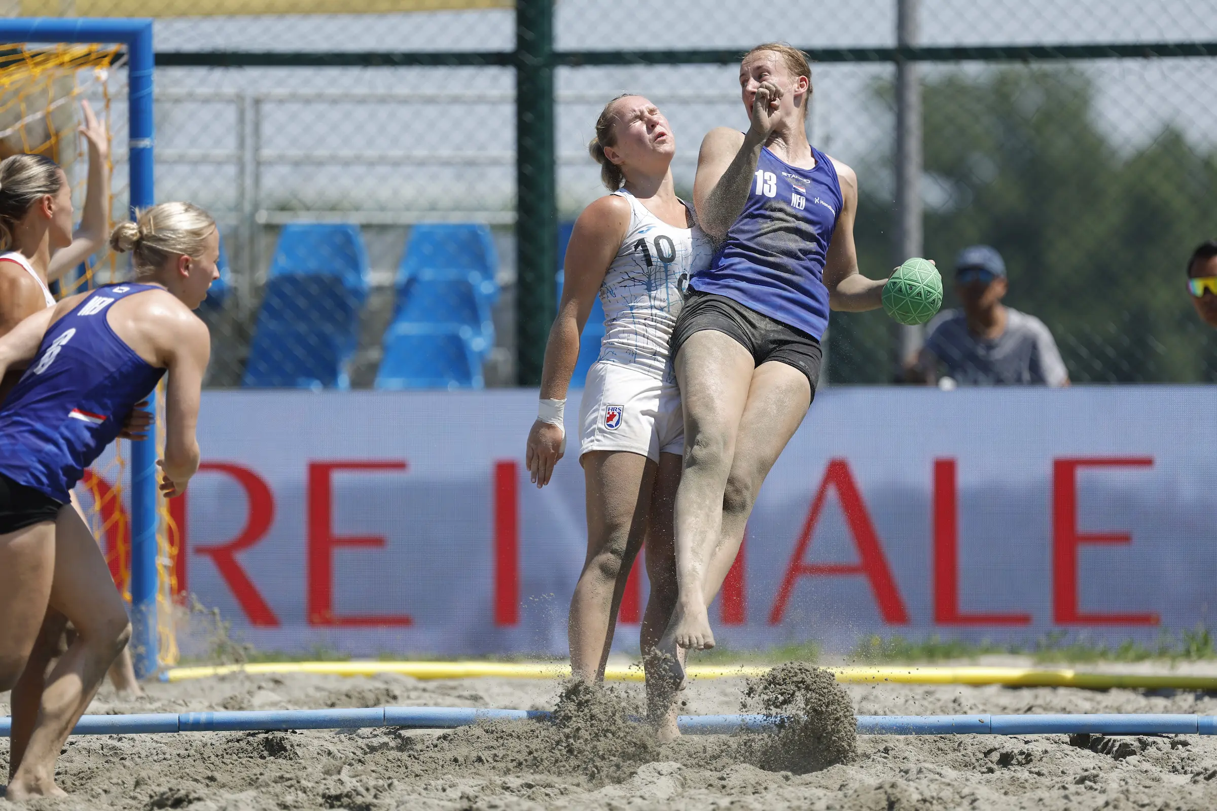 Two athletes compete in a dynamic sand volleyball match, with one player leaping to strike the ball while the other defends.