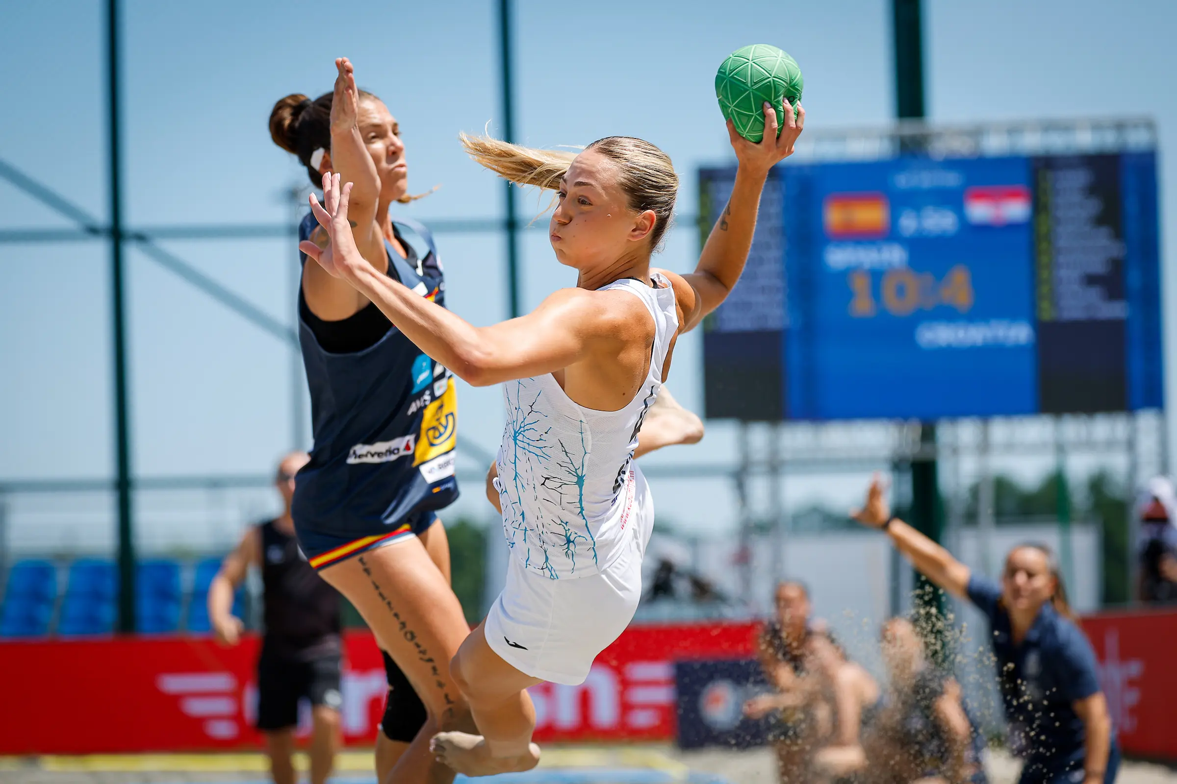 RUKOMET NA PIJESKU An athlete in a white uniform leaps to pass a green ball while being defended by an opponent on a sunny outdoor court.